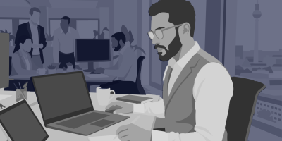 Ein bärtiger Mann mit Brille und Weste arbeitet im Großraumbüro an einem Laptop; im Hintergrund Kolleginnen und Kollegen sowie der Berliner Fernsehturm hinter dem Fenster.
