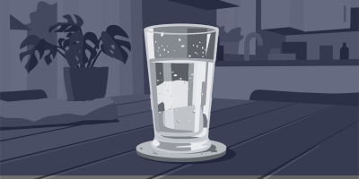 Ein bis zur Hälfte mit Wasser gefülltes Trinkglas steht auf einem Untersetzer auf einem Holztisch. Im Hintergrund eine Zimmerpflanze und eine angedeutete Küche.