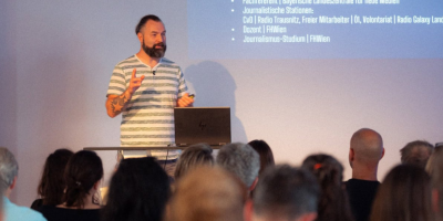 Marcel Tuljus steht mit Laptop an einem Stehpult und spricht zu einem Publikum, das von hinten zu sehen ist.