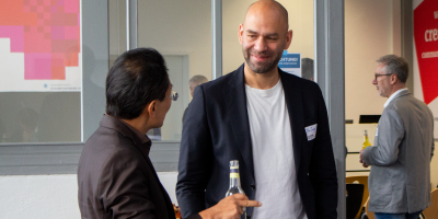 Chan-jo Jun und Jim Sengl stehen bei einer Veranstaltung mit Getränkeflaschen in der Hand einander zugewandt und unterhalten sich lächelnd; im Hintergrund weitere Gäste.